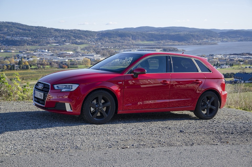 Audi A3 Sportback 8V rojo de tres cuartos delantero, aparcado sobre grava con un fiordo y colinas al fondo