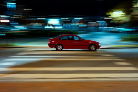 Coche antiguo de color rojo cruzando un paso de peatones de noche, con las luces de la ciudad desenfocadas al fondo