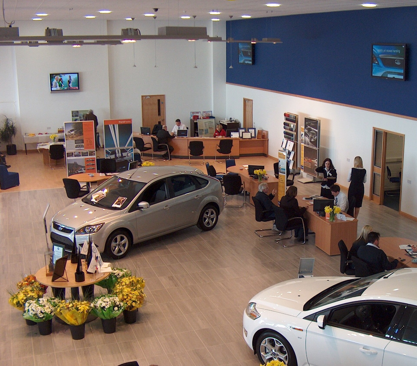 Sala de exposición de un concesionario con dos coches expuestos y comerciales atendiendo a clientes en las mesas