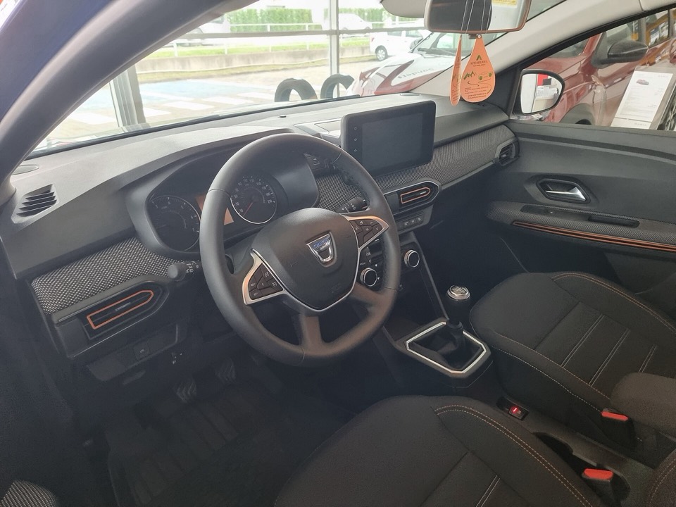 Puesto de conducción de un Dacia Sandero III con pantalla central, cambio manual y molduras naranjas