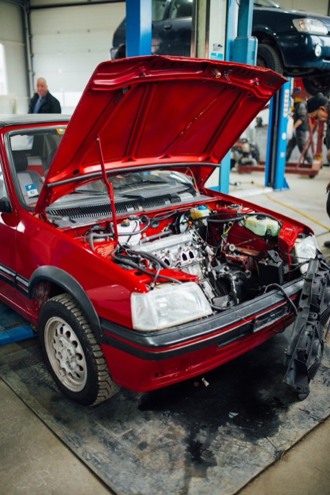 Peugeot 205 rojo con el capó abierto en un taller, con otro coche sobre el elevador al fondo