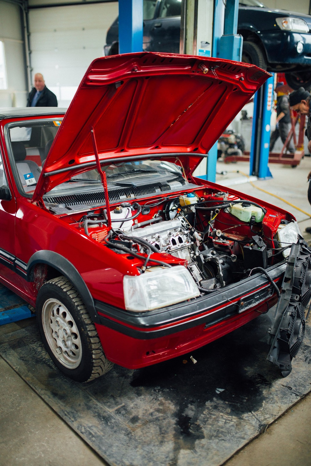 Peugeot 205 rojo con el capó abierto en un taller, con otro coche sobre el elevador al fondo
