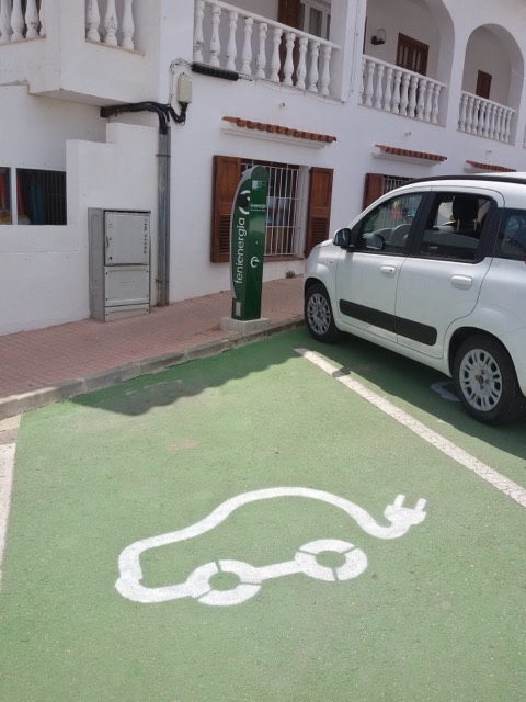 Plaza de aparcamiento española pintada de verde con el pictograma de coche eléctrico y un poste de recarga junto a un utilitario blanco