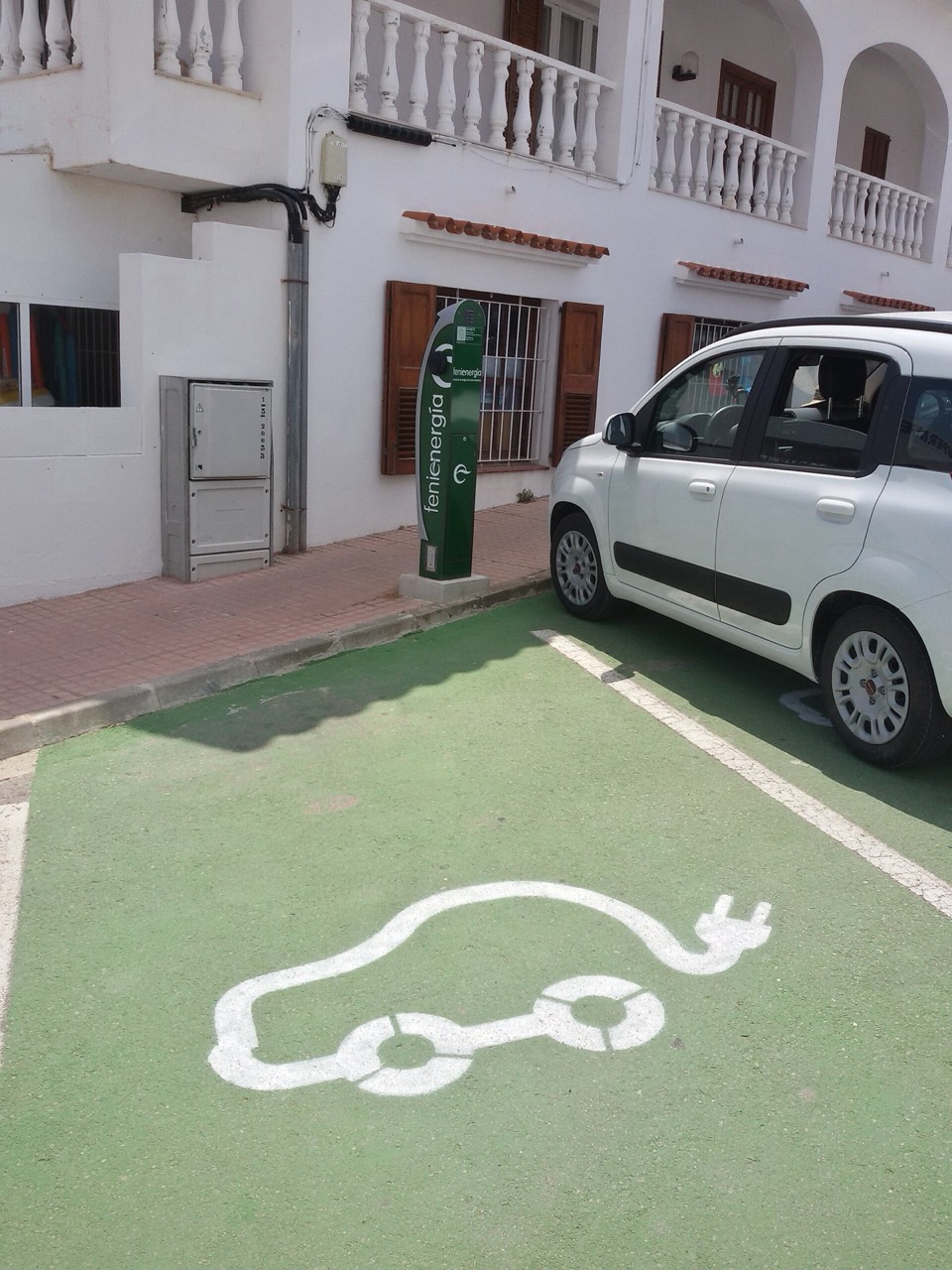 Plaza de aparcamiento española pintada de verde con el pictograma de coche eléctrico y un poste de recarga junto a un utilitario blanco