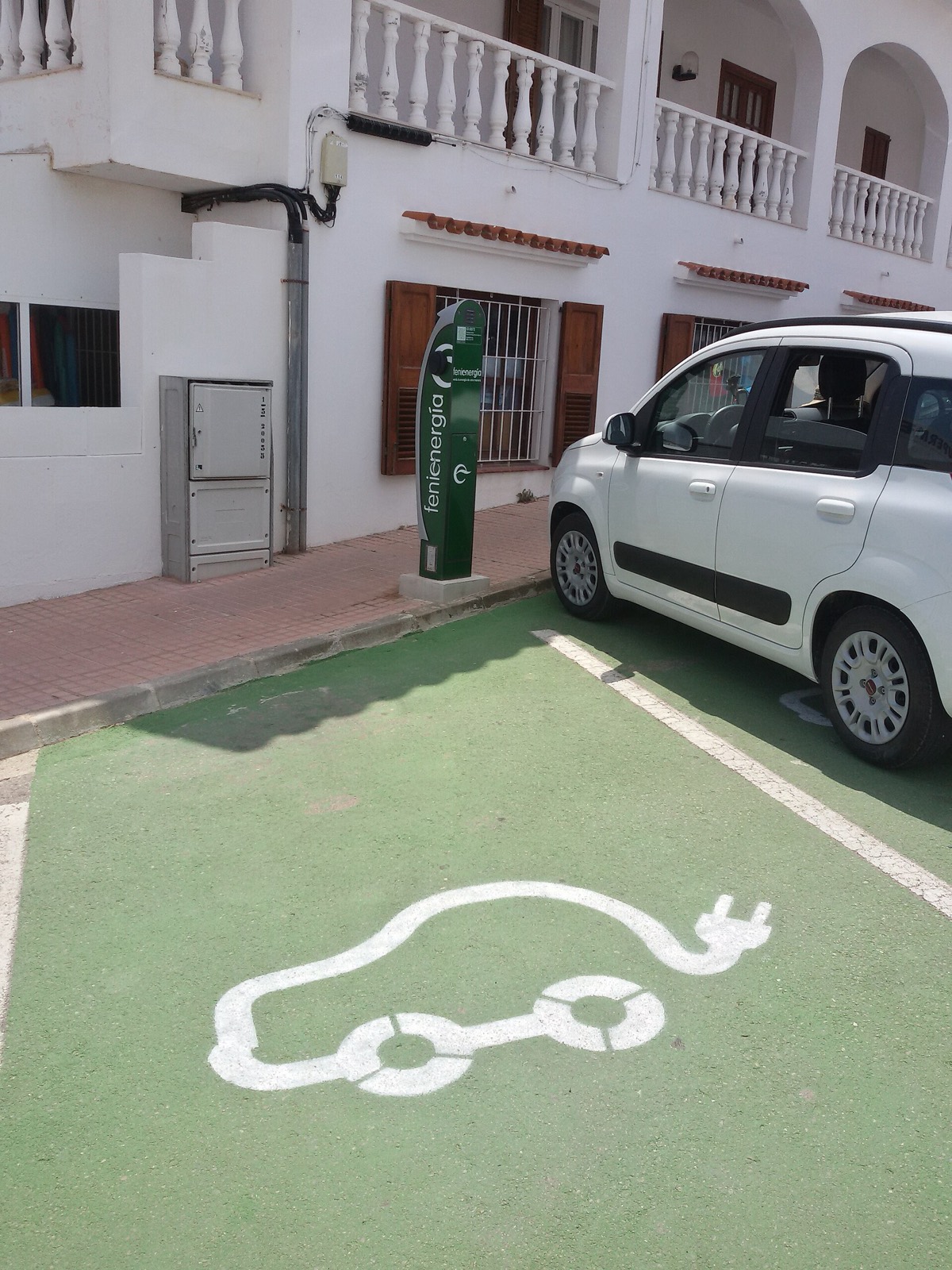 Plaza de aparcamiento española pintada de verde con el pictograma de coche eléctrico y un poste de recarga junto a un utilitario blanco
